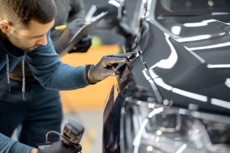 A person checks a car’s engine oil dipstick in a residential driveway on a sunny day.